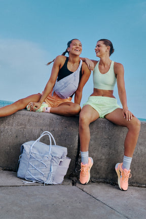 Two women in athletic wear smiling, sitting on a ledge with a bag.